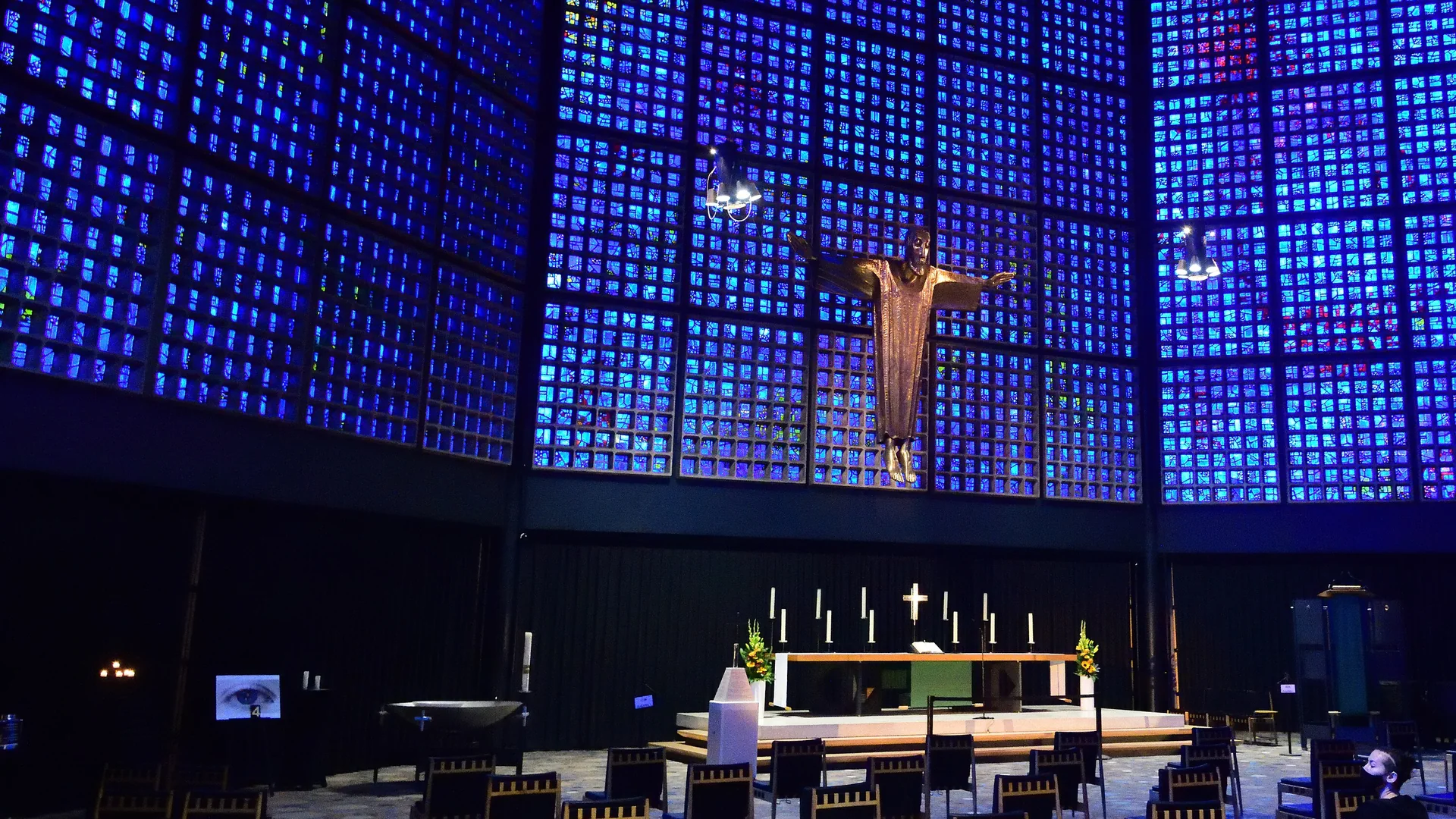 Das Kirchenschiff der Kaiser-Wilhelm-Gedächtniskirche in Berlin: Wände aus kleinen blauen Glasfenstern erheben sich um eine vergoldete Christusfigur über dem Altar, auf dem Boden stehen Holzstühle.