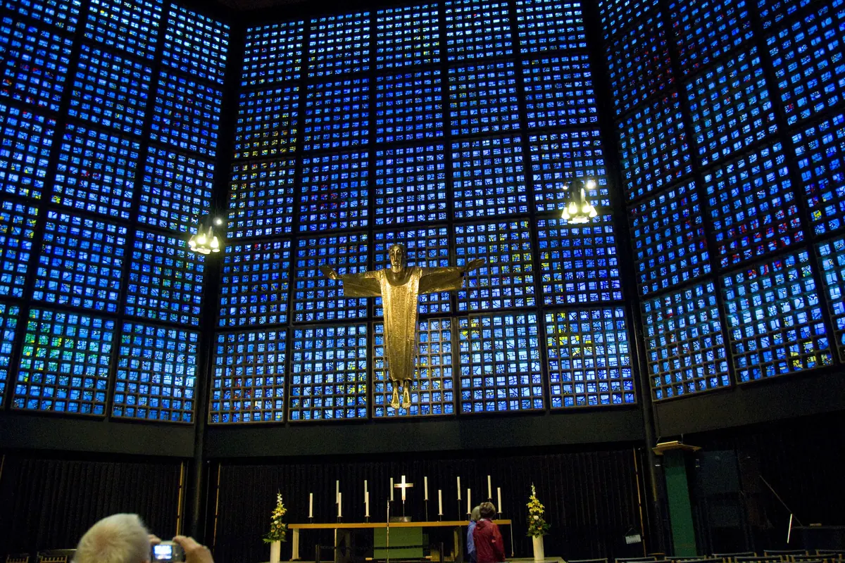 Die blaue Glaswand des Kirchenschiffs der Gedächtniskirche, die sich hinter dem Altar erhebt, mit einer vergoldeten Christusfigur in der Mitte der Wand und Reihen von Holzstühlen davor