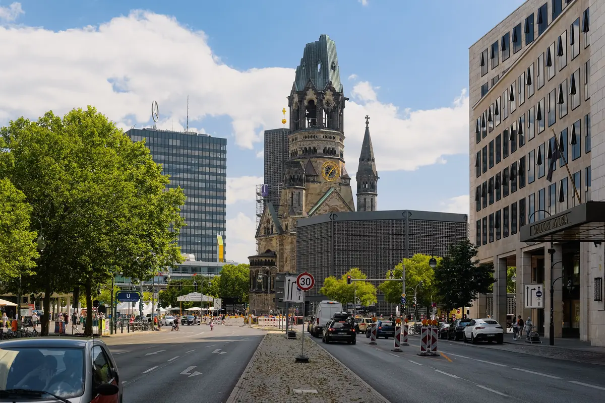 Der Turm der Gedächtniskirche, gesehen eine Berliner Straße hinunter bei Tageslicht, die zerbrochene neuromanische Turmspitze neben dem niedrigen modernen Achteck und Turm sowie Stadtgebäude auf beiden Seiten