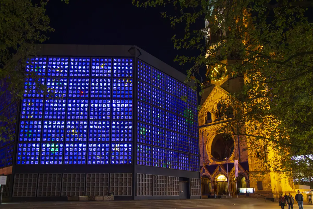 Die neue Gedächtniskirche bei Nacht, ihre blauen Glaswände leuchten neben dem angestrahlten Sandsteinturm der alten Kirche und einer beleuchteten Straße davor